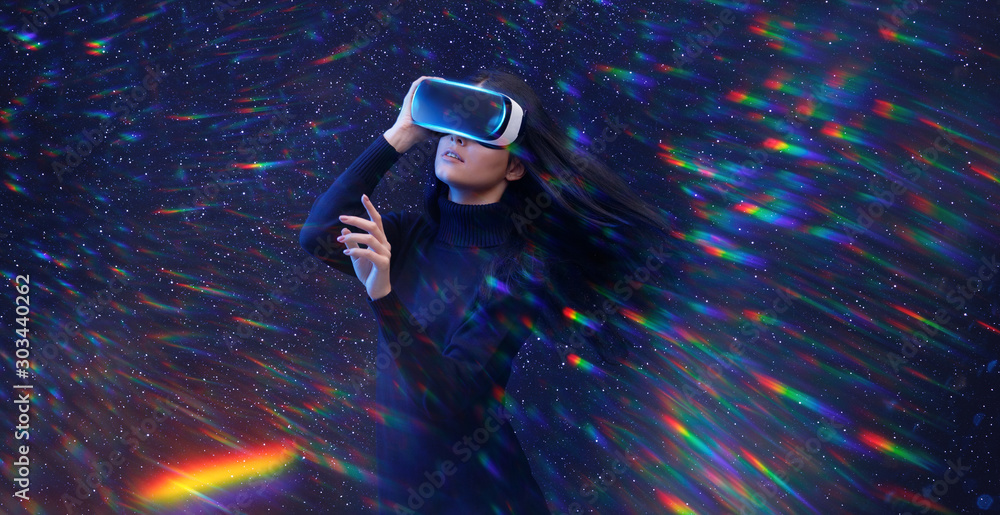 Beautiful young woman using VR helmet while touching air in colorful  rainbow neon lights. Girl in glasses of virtual reality with blue backlight  with flowing hair over dark magic universe background. Stock