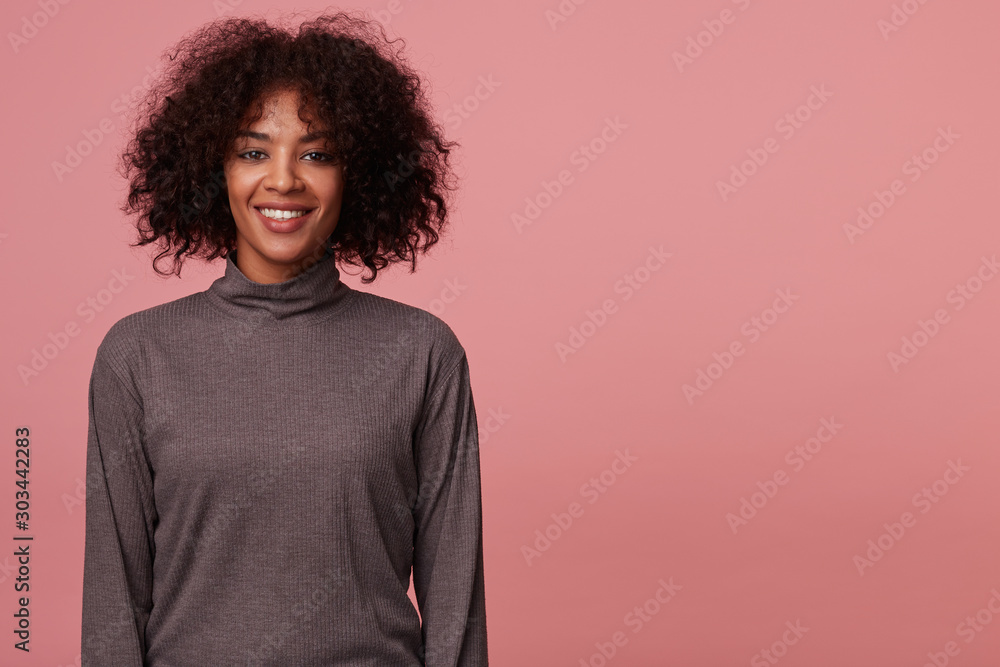 Cheerful pretty young curly brunette female with dark skin wearing casual hairstyle, keeping hands along her body while standing over pink background in grey poloneck Cheerful pretty young curly brunette female with dark skin wearing casual hairstyle, keeping hands along her body while standing over pink background in grey poloneck