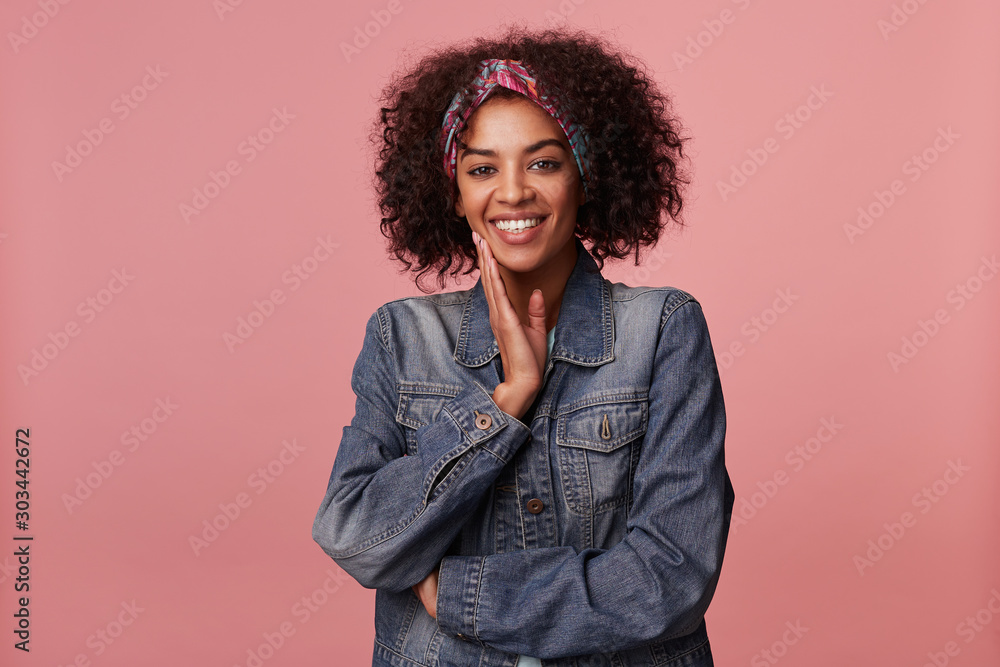 Joyful attractive young dark skinned lady with short curly brown hair wearing colorful headband and touching gently her face with raised palm, smiling widely to camera over pink background Joyful attractive young dark skinned lady with short curly brown hair wearing colorful headband and touching gently her face with raised palm, smiling widely to camera over pink background