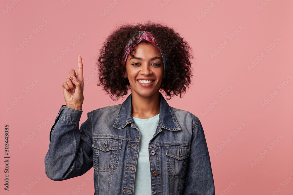 Pleased positive young curly brunette lady with dark skin wearing casual hairstyle, smiling widely to camera and raising up index finger, wearing mint t-shirt and jeans coat over pink background Pleased positive young curly brunette lady with dark skin wearing casual hairstyle, smiling widely to camera and raising up index finger, wearing mint t-shirt and jeans coat over pink background