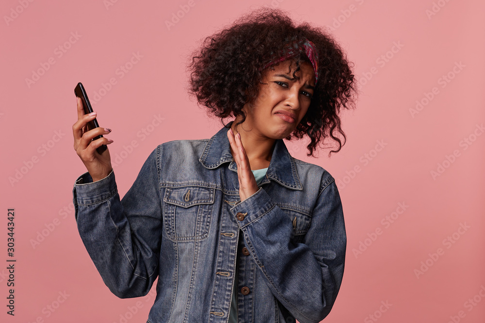 Portrait of displeased young curly brunette female with dark skin moving away from herself handset and looking to camera with pout, dressed in casual wear while standing over pink background Portrait of displeased young curly brunette female with dark skin moving away from herself handset and looking to camera with pout, dressed in casual wear while standing over pink background