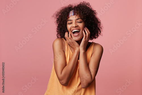 Pleasant looking happy young curly brunette female with dark skin keeping hands on cheeks and smiling widely with closed eyes, dressed in light orange top and colorful headband over pink background