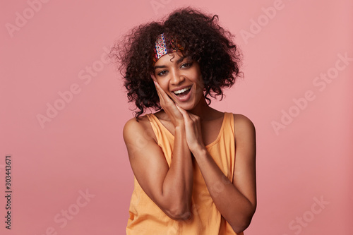 Happy young curly brunette lady with dark skin wearing colorful headband and light orange top while standing over pink background, smiling cheerfully to camera and keeping hands under her cheek