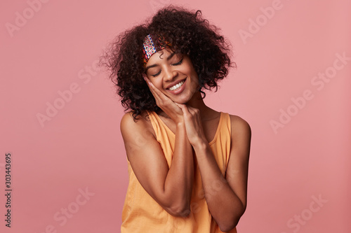 Sleepy pretty dark skinned curly brunette female with short haircut leaning her cheek on raised hands and keeping eyes closed, smiling sincerely while posing over pink background