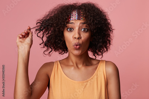 Portrait of charming young curly brunette female with dark skin looking surprisedly at camera and curling lips, playing with her short hair while standing over pink background in casual wear