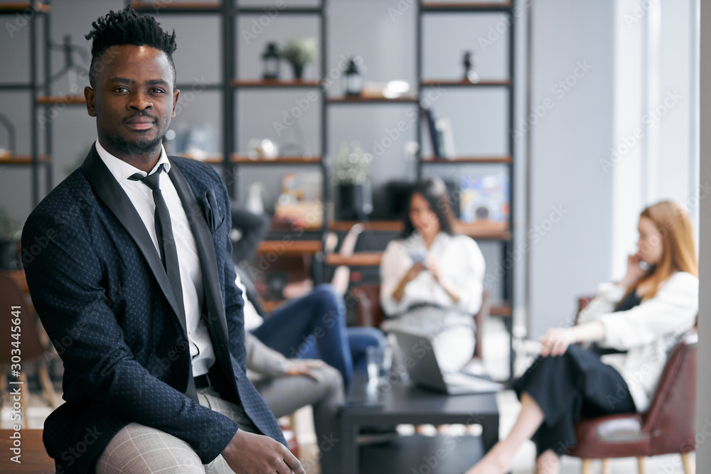 Good-looking african man wearing black suit stand posing in modern office and look at camera. Colleagues background Good-looking african man wearing black suit stand posing in modern office and look at camera. Colleagues background