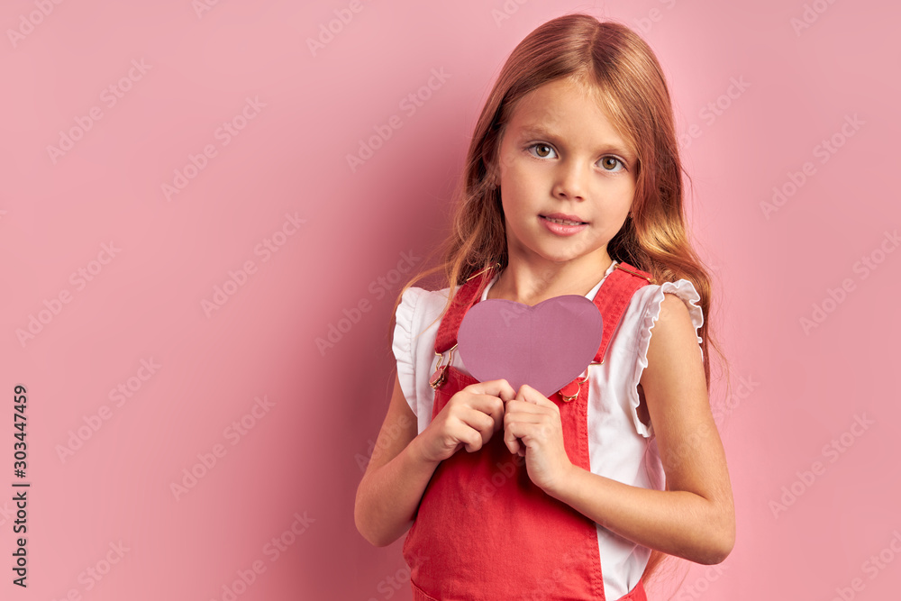 Portrait of beautiful girl wearing red overalls and holding purple cardboard heart, having golden long hair. Look at camera isolated over pink background Portrait of beautiful girl wearing red overalls and holding purple cardboard heart, having golden long hair. Look at camera isolated over pink background