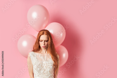 Young female with auburn hair stand sadly looking at floor, sadness. Isolated over pink background