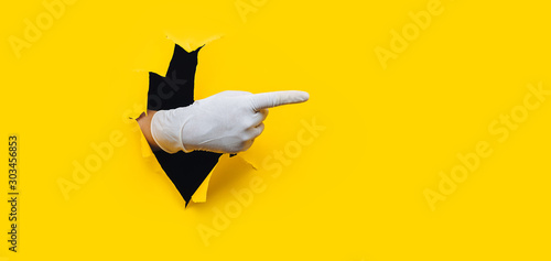 The forefinger points in white medical glove to the right side. Yellow paper background with torn hole. Place for advertising. Copy space. photo