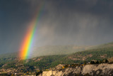 Vibrant rainbow shining through storm clouds and falling rain on forested hillside