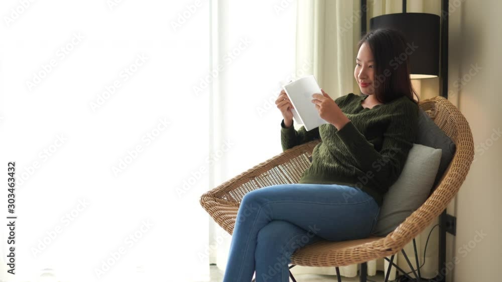 Young Asian woman wearing casual jeans and a green sweater is sitting in a rattan chair thumbing through and reading a small book. Young Asian woman wearing casual jeans and a green sweater is sitting in a rattan chair thumbing through and reading a small book.