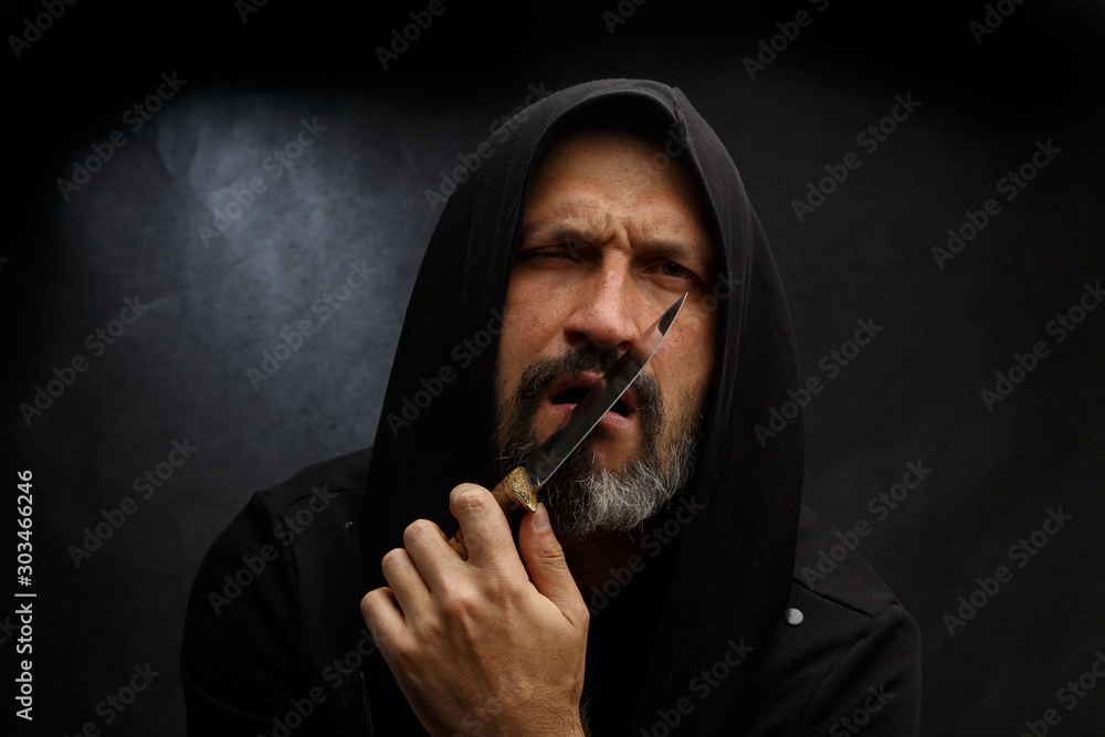 Portrait of a bald man with a beard in a black hood with a knife on a dirty gray background. Maniac. Portrait of a bald man with a beard in a black hood with a knife on a dirty gray background. Maniac.