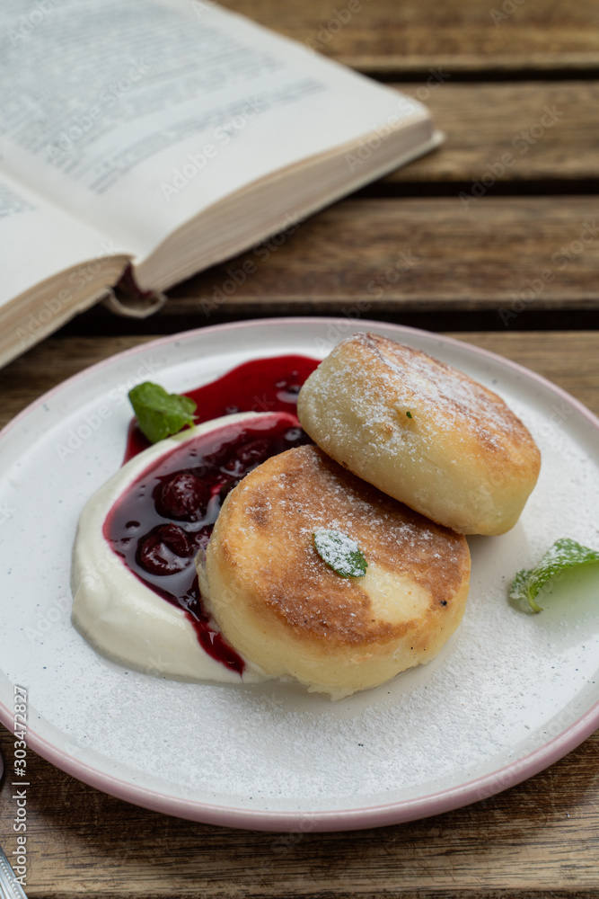 Puffy cheese pancakes with sour cream and cherry sauce on a wooden table with book