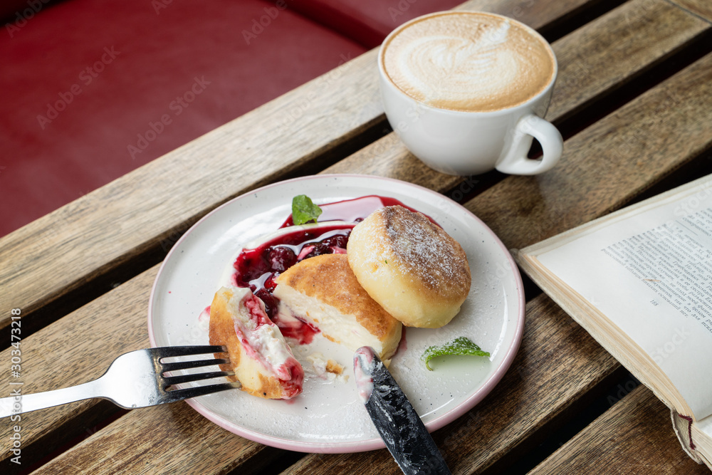 Cheese pancakes with sour cream and cherry sauce and cappuccino on a wooden table with book Cheese pancakes with sour cream and cherry sauce and cappuccino on a wooden table with book