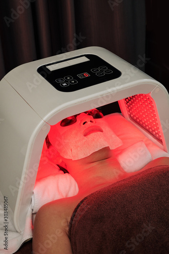 Woman having color light therapy and lying in sheet mask under red light lamp