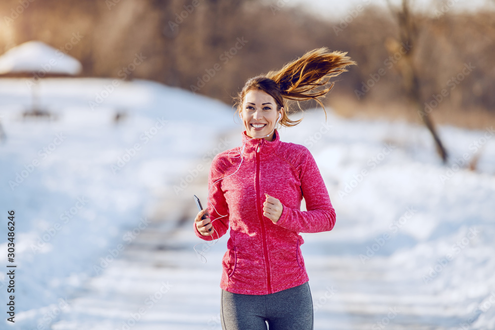 Φωτογραφία Smiling caucasian brunette in sportswear, with earphones in ears and with smart phone in hand running in nature