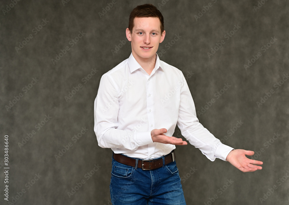 Concept of a pleasant young man manager talking to the camera. Photo Portrait of a secretary guy on a gray background in blue jeans and a white shirt in various poses. Concept of a pleasant young man manager talking to the camera. Photo Portrait of a secretary guy on a gray background in blue jeans and a white shirt in various poses.