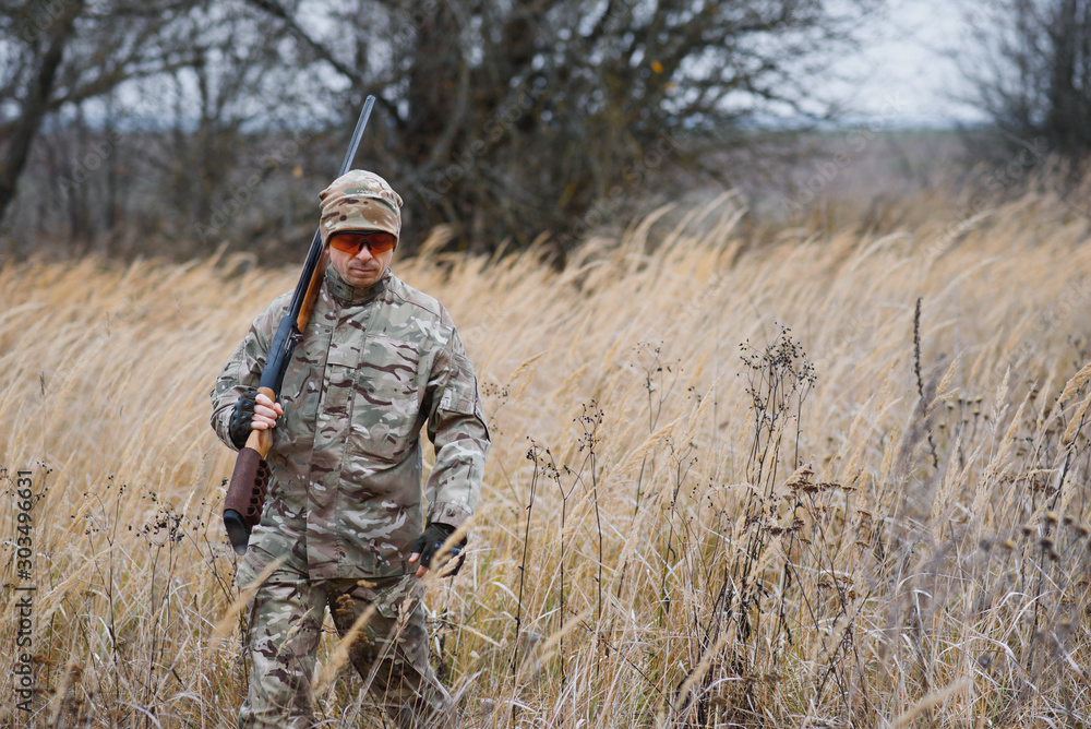 A male hunter with a gun while sitting takes aim at a forest. The concept of a successful hunt, an experienced hunter. Hunting the autumn season. The hunter has a rifle and a hunting uniform.