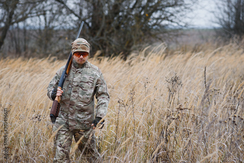 A male hunter with a gun while sitting takes aim at a forest. The concept of a successful hunt  an experienced hunter. Hunting the autumn season. The hunter has a rifle and a hunting uniform.