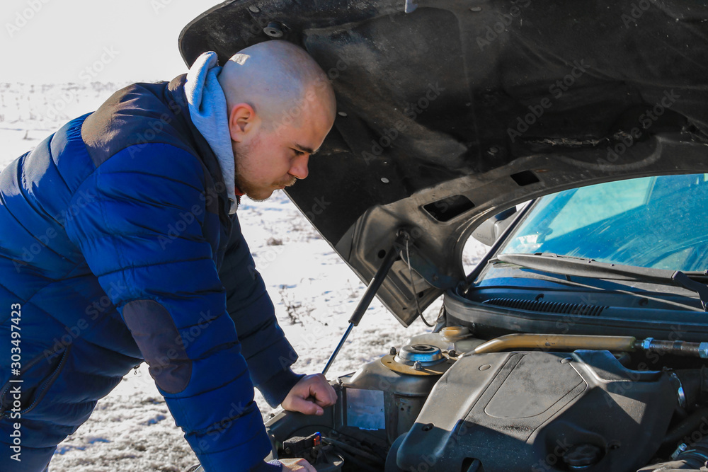 Young man looking under the hood of broken car in cold winter day Young man looking under the hood of broken car in cold winter day