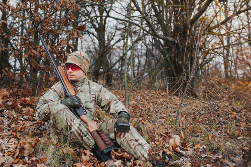 A male hunter with a gun while sitting takes aim at a forest. The concept of a successful hunt, an experienced hunter. Hunting the autumn season. The hunter has a rifle and a hunting uniform.