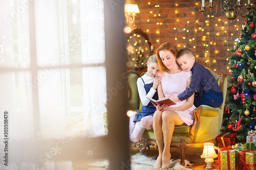 Happy mother reads fairy tales to children while sitting on a sofa near a festive Christmas tree