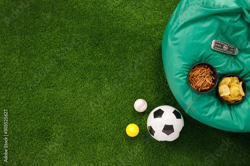 Soccer ball on a green field and ottoman for a fan with snacks and a TV remote control. The apartment was lying. The concept of football matches.