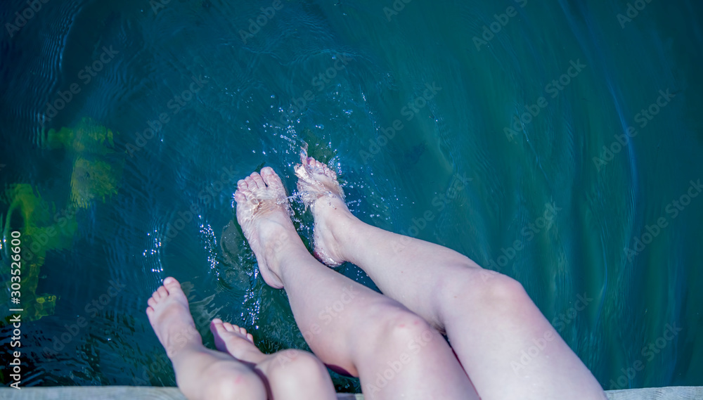 Fototapeta premium Top view of mother and her daughter have fun on pier splashing their legs into water of pond.