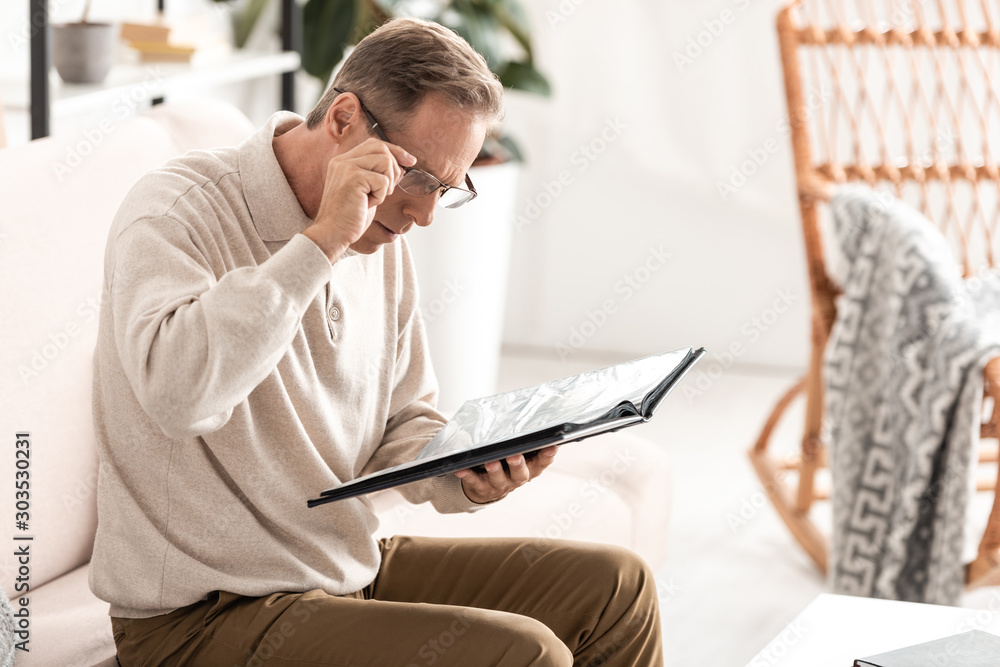 senior man touching glasses and holding photo album while sitting on sofa senior man touching glasses and holding photo album while sitting on sofa