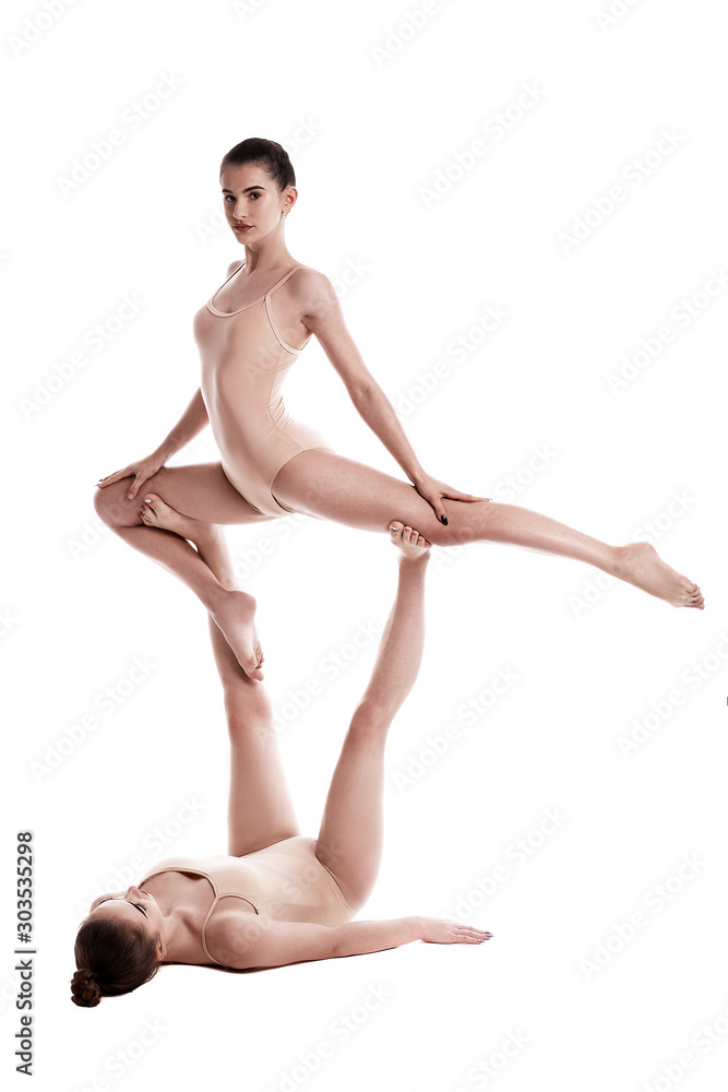 Two flexible girls gymnasts in beige leotards are performing exercises upside down using support and posing isolated on white background. Close-up. Two flexible girls gymnasts in beige leotards are performing exercises upside down using support and posing isolated on white background. Close-up.