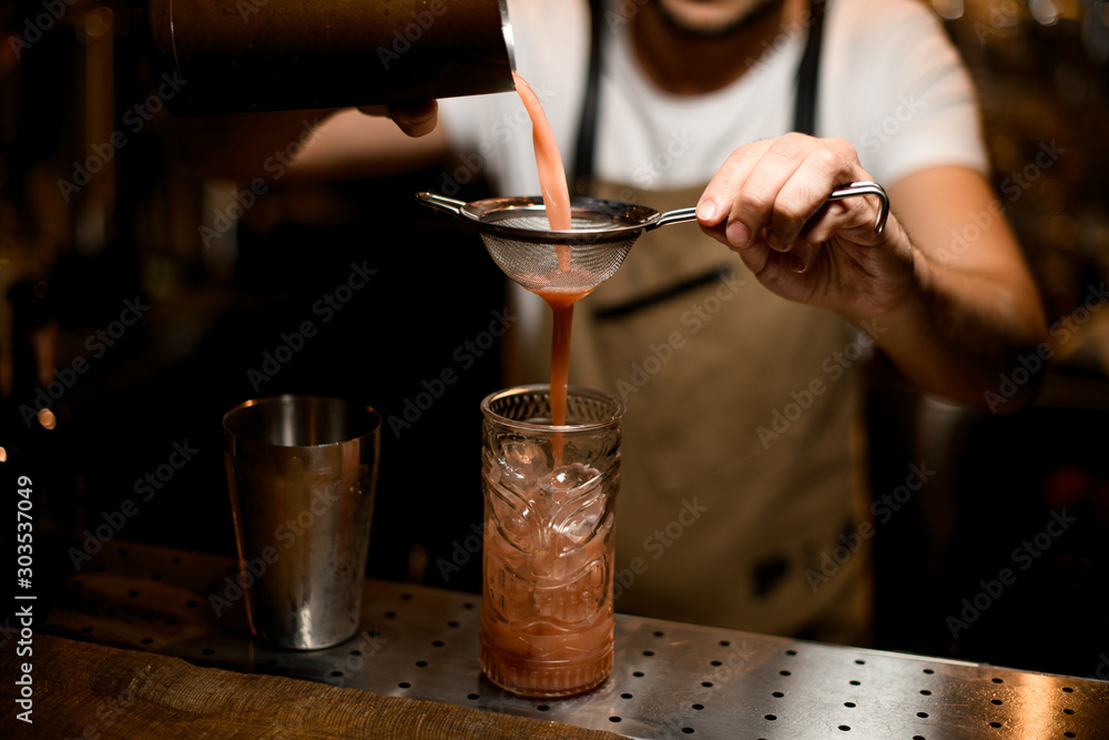 Professional male bartender pouring a red alcoholic drink from steel shaker through the sieve