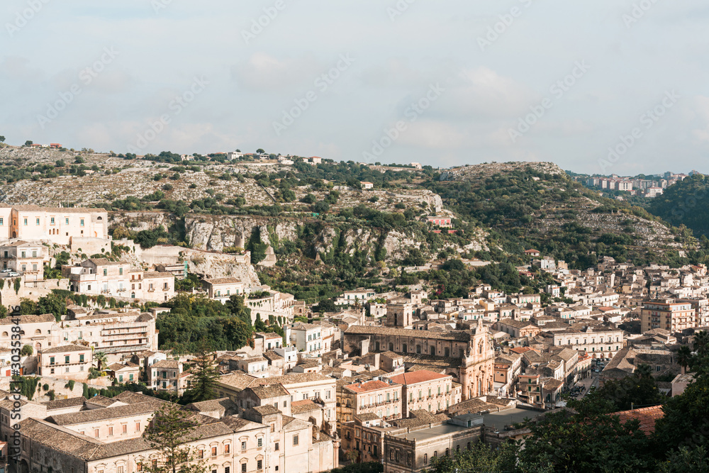 sunlight on small houses near green trees against sky with clouds in ragusa, italy