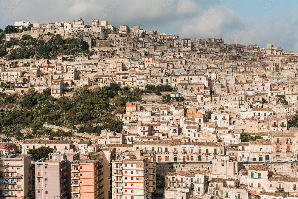 sunlight on houses near green trees against sky with clouds in ragusa, italy