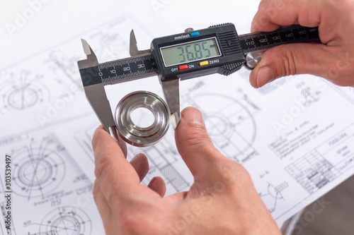 Hands of an engineer measures a metal part with a digital vernier caliper against the background of technical drawings. Quality control of parts machined on a lathe.