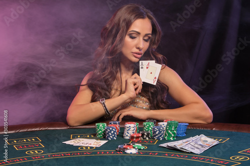 Girl playing poker, casino. Showing cards, sitting at table with stacks of chips and money. Black, smoke background, colorful backlights. Close-up. photo