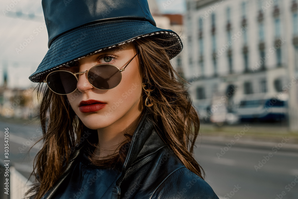 Outdoor close up fashion portrait of young elegant model, woman wearing  trendy faux leather bucket hat, round sunglasses, earrings, posing in  street of European city. Copy, empty space for text Stock Photo