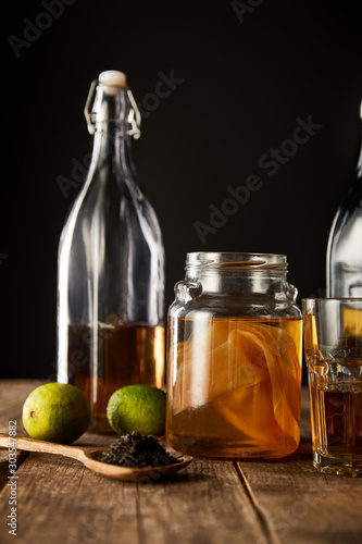 glass jar with kombucha near lime, spice and bottle on wooden table isolated on black