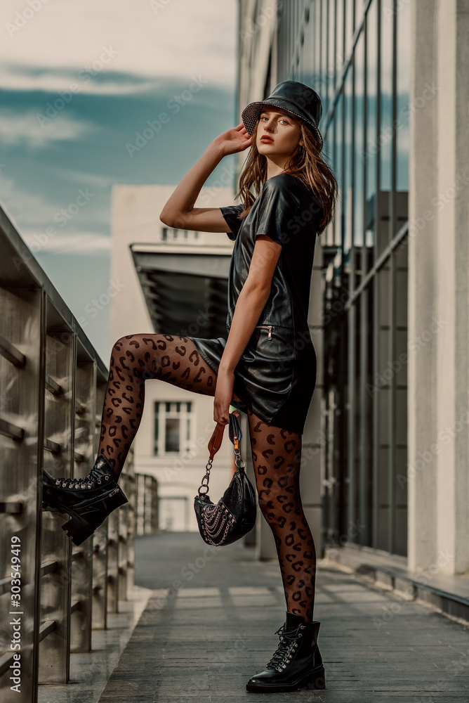 Outdoor full body street fashion portrait of young elegant model, woman  wearing trendy leather bucket hat, short dress, black leopard print tights, lace  up ankle boots, holding small stylish handbag Stock Photo