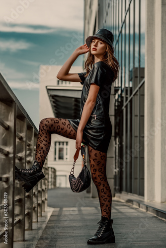 Outdoor full body street fashion portrait of young elegant model, woman  wearing trendy leather bucket hat, short dress, black leopard print tights,  lace up ankle boots, holding small stylish handbag Stock Photo