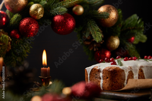 traditional Christmas cake with cranberry near Christmas wreath with baubles and candle on wooden table isolated on black