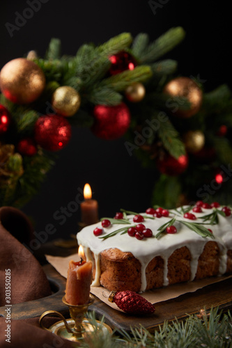 selective focus of traditional Christmas cake with cranberry near Christmas wreath with baubles and burning candles on wooden table isolated on black