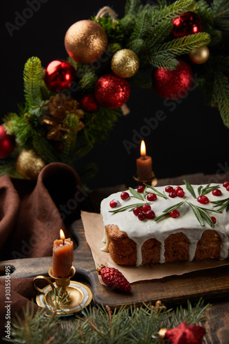 traditional Christmas cake with cranberry near Christmas wreath with baubles and candles on wooden table isolated on black