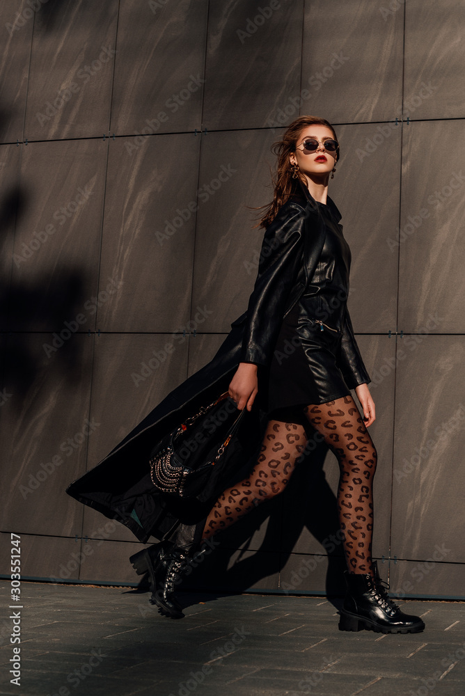 Outdoor full-length fashion portrait of young confident woman wearing total  black leather outfit, leopard print tights, lace up boots, holding small  bag, walking in city street, grey urban background Stock Photo