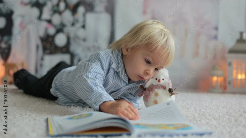 Blond toddler child, boy, reading a book at home, lying on the floor with a toy Blond toddler child, boy, reading a book at home, lying on the floor with a toy