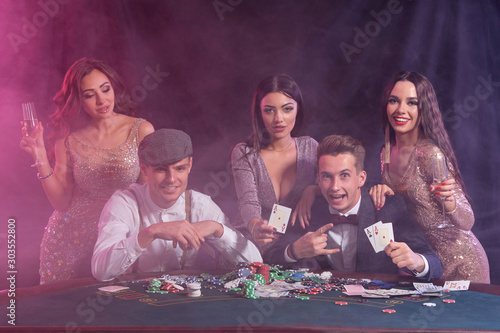Friends playing poker at casino, at table with stacks of chips, money, cards on it. Celebrating win, smiling. Black, smoke background. Close-up.