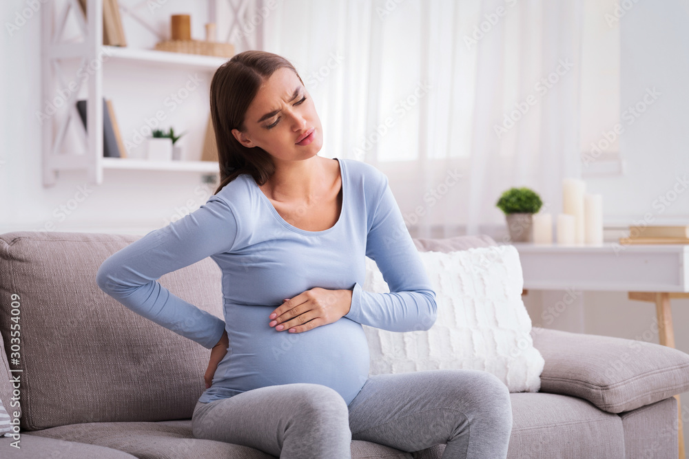 Obraz premium Pregnant Lady Having Backache Massaging Back Sitting On Sofa Indoor