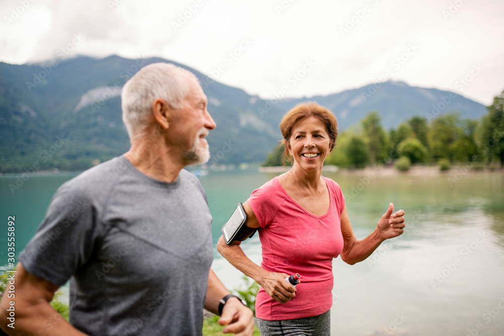 Senior pensioner couple with smartphone running by lake in nature.