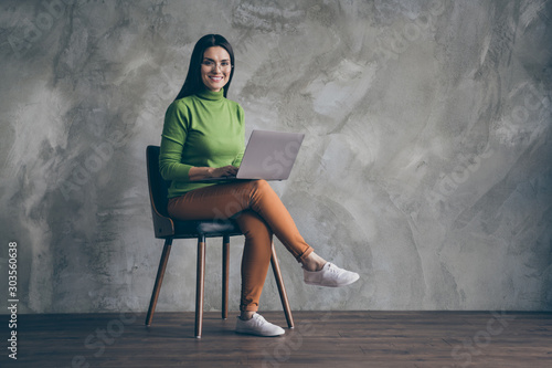 Turned full length body size photo of cheerful girlfriend sitting in chair working with laptop confident developing new project to present isolated grey wall color concrete background