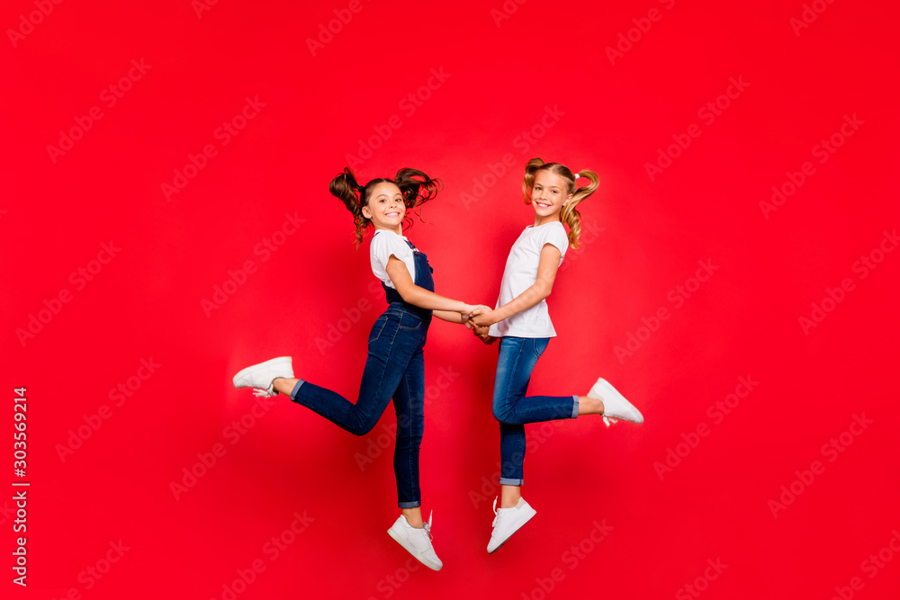 Full length profile side photo of two small kids having x-mas weekends jump hold hands wear white t-shirt denim jeans overalls sneakers isolated over shine color background