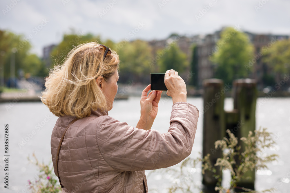 An elderly woman photographs the sights of the city on a mobile phone, walking through the streets of the summer city. Concept of active lifestyle of elderly people.